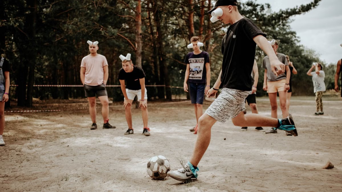Group of teenagers wearing masks playing a soccer game in a sandy outdoor setting.